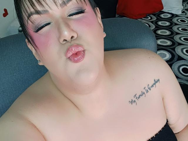 GiiannaMorrys with pouty pink lips, short black hair, and pink eyeshadow, showing off her bare shoulders and tattoo on her right chest, sitting on a gray couch.