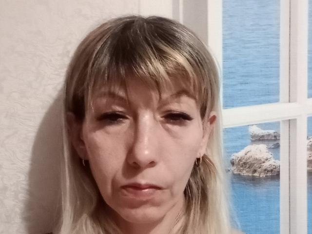 Nerpenok has blonde hair with bangs, slightly closed eyes, and small earrings. Her face shows a subtle, almost unamused, expression. A window with blue water is visible.