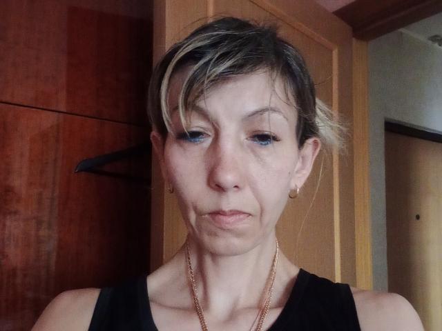 Nerpenok tiene el cabello corto con mechas rubias, ojos azules y una expresión pensativa. Lleva una camiseta negra sin mangas, aretes dorados y un collar dorado. Su rostro muestra arrugas sutiles..