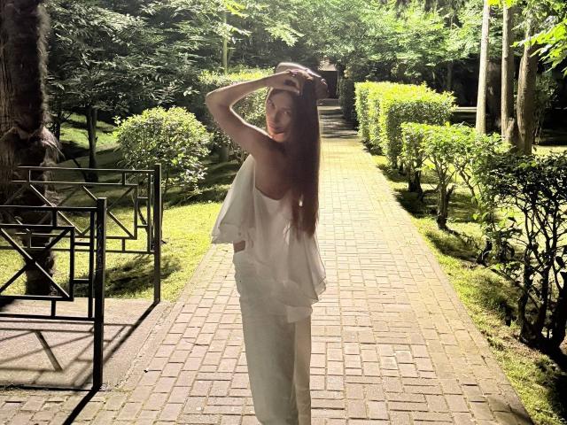 Adriana Nalley mit langen braunen Haaren, einem weißen, ärmellosen Kleid und weißen Hosen, steht auf einem Ziegelweg, eine Hand im Haar, und blickt mit einem leichten Lächeln zurück..