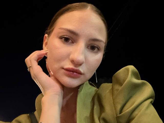 AdrianaNalley met volgezwollen lippen, een groene jurk, een hand die haar wang aanraakt, bruine ogen en glad, naar achteren getrokken haar, die recht in de camera kijkt, tegen een donkere achtergrond..