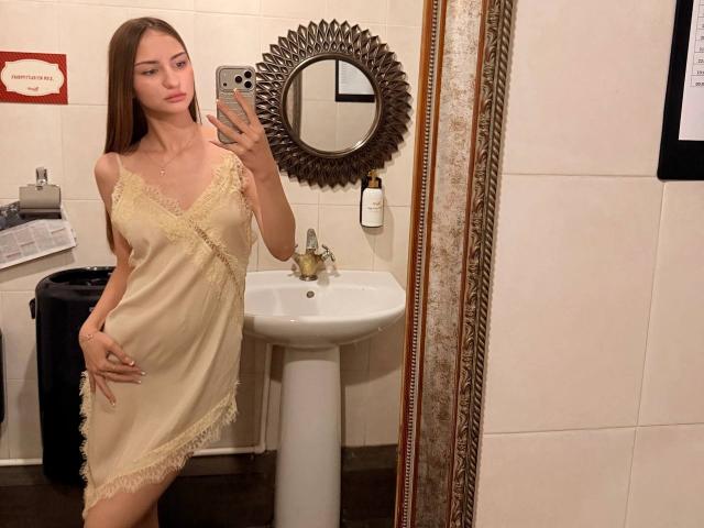 Adriana Nalley, vêtue d'une nuisette soyeuse beige avec des détails en dentelle, se tient dans une salle de bain et prend un selfie devant le miroir, sa main droite posée sur sa hanche, mettant en valeur sa petite poitrine..