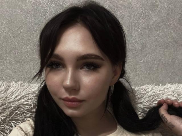 LawandaMuzzy with dark pigtails, holding one side, has light skin, thick eyebrows, long eyelashes, and pink lips, wearing a beige top, looking at the camera with a slight smile.