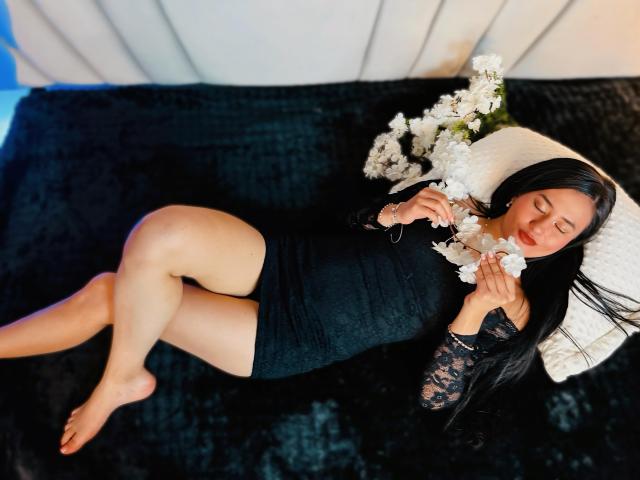 SarahMoonOne, allongée sur une couverture noire texturée, portant une robe noire en dentelle, tenant des fleurs blanches, les yeux fermés, de longs cheveux noirs, des jambes nues, un oreiller blanc, un éclairage doux, une pose détendue..