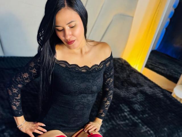 SarahMoon, une femme aux longs cheveux noirs, portant une robe noire en dentelle à épaules dénudées, assise sur un lit noir, les mains posées sur ses cuisses, regardant vers le bas, avec des talons hauts rouges devant elle..