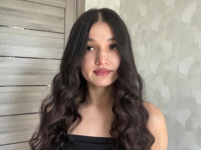 YoulandaCarlough with long, wavy black hair, brown eyes, nose ring, and pinkish lipstick, wearing a black strapless top, showing her boobs slightly.
