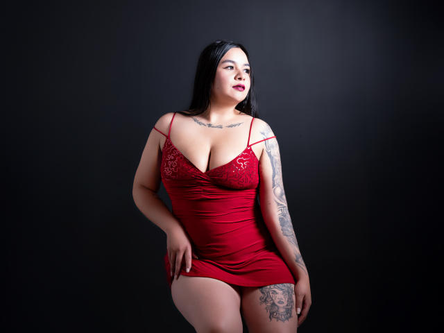 EveWillis with big tits in a red lace dress, black hair, tattoos, looking to the side, dark background, hand on thigh, standing, sexy, confident, dark red lipstick.