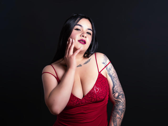 EveWillis with black hair, red lace dress, big boobs, tattooed arm, hand on cheek, red lipstick, dark background, sexy, confident.