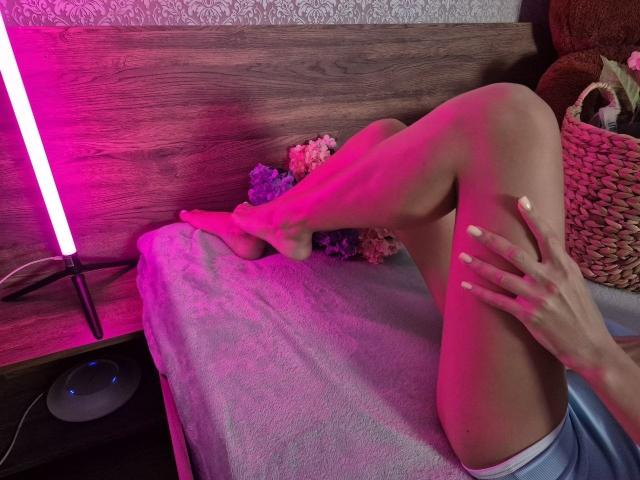 As pernas de TenerliRize estão cruzadas em uma cama com uma luz neon rosa, mostrando pele lisa e unhas brancas. Uma cesta de vime com flores está à direita..