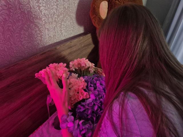 TenerliRize, avec de longs cheveux bruns, des mèches roses et violettes, tenant des fleurs roses et violettes, appuyée contre une tête de lit en bois avec un ours en peluche marron accroché au mur..