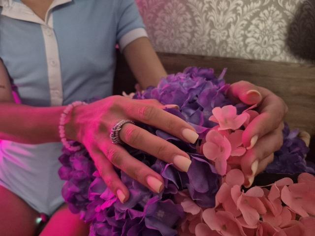 As mãos da TenerliRize, com unhas cor-de-rosa, usando um anel, segurando pétalas de flores roxas e cor-de-rosa, reflexo de luz rosa, camisa azul, superfície de madeira, papel de parede estampado ao fundo..