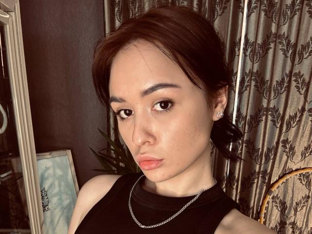 AnnaBauknight with pouty lips, brown eyes, and dark brown hair, wearing a black sleeveless top and silver chain, has small earrings and a slight tan, in a bedroom.