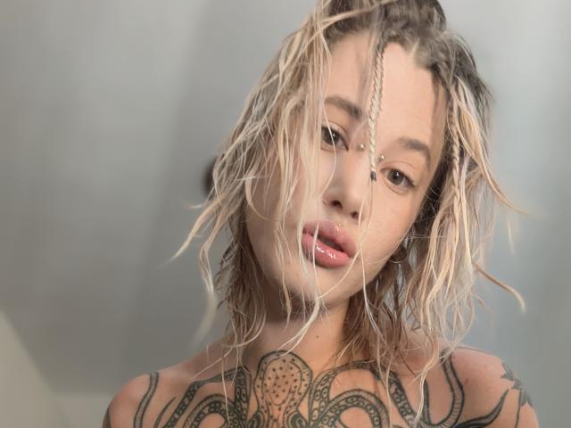 MadameSandra, com cabelo loiro úmido, tatuagens no peito e um piercing no nariz, olhando para a câmera com lábios rosados e brilhantes. Seus seios estão parcialmente visíveis..