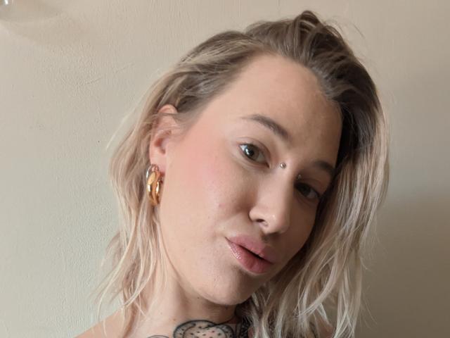 MadameSandra tiene el cabello rubio y ondulado, un arete de aro dorado, una marca de belleza en la cara y un tatuaje debajo del cuello. Está sonriendo con los labios rosados ligeramente entreabiertos..