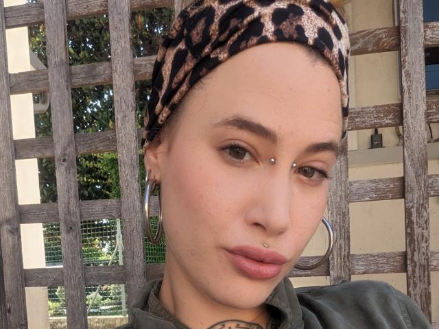 MadameSandra indossa una sciarpa con motivo leopardato, orecchini a cerchio, un piercing al naso, un piercing al labbro e un piercing al setto, e una maglietta verde, che mette in mostra le sue labbra carnose e provocanti..