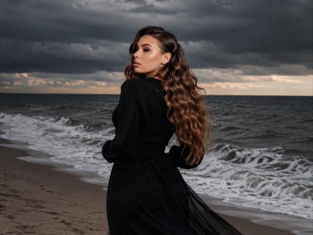 Niferttiti, con el pelo largo y ondulado de color marrón, vestida con un vestido negro, está de pie en una playa con cielos oscuros y nublados y olas del océano, mirando por encima del hombro, con una expresión seria..