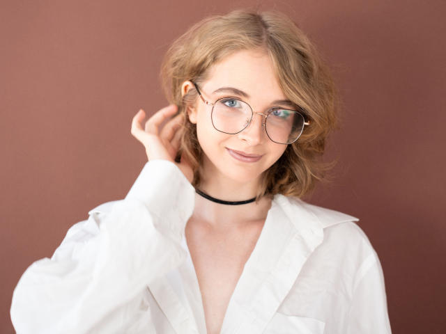 PennyDelight con capelli biondi, occhiali, camicia bianca, collana nera, che sorride, con una mano tra i capelli, occhi azzurri, carina, luce soffusa, trucco leggero, leggero rossore, rilassata, casual, espressione dolce..