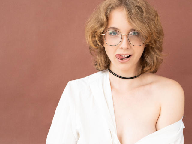 PennyDelight with curly blonde hair, glasses, black choker, and white shirt partially revealing her tits, sticking her tongue out. Soft brown background.