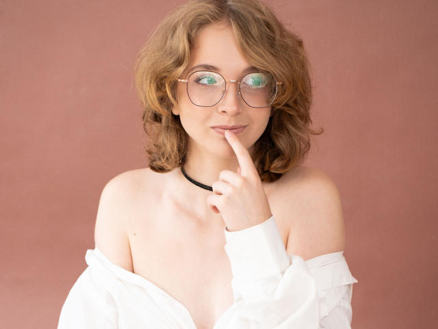PennyDelight, con cabello castaño ondulado, gafas y un collar negro, vestida con una camisa blanca con un hombro descubierto, tiene un dedo en los labios, mostrando una expresión linda y pensativa..