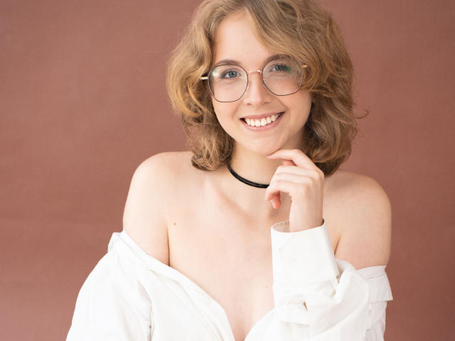 PennyDelight with curly brown hair, glasses, smiling, white shirt off shoulders, black choker, hand near mouth, showing her bare tits, pinkish background.