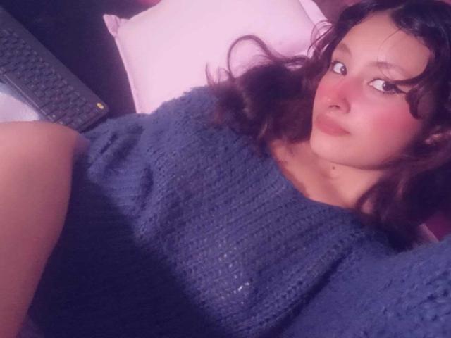 ValentinaRoseX with wavy brown hair, wearing a blue knitted sweater, lying down, looking at the camera, her tits slightly visible under the sweater, pink pillow in background.