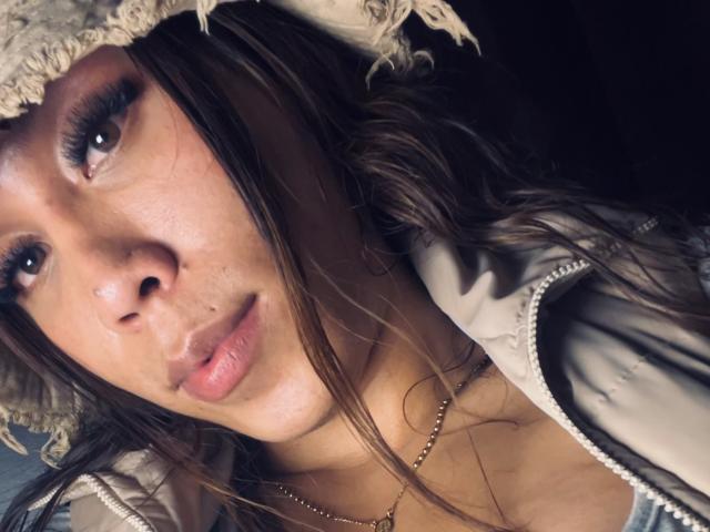 Geraldine Suan, met lang bruin haar, draagt een beige hoed en een dikke jas. Ze heeft bruine ogen, volle, roze lippen en een kleine neus, en draagt een gouden ketting..