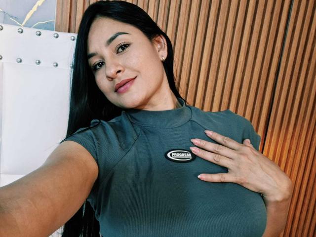 Exotische Latina met lang zwart haar, die een grijze geribbelde top draagt, met haar hand op haar borst, waarbij kleine borsten zichtbaar zijn, een subtiele glimlach, een houten achtergrond, een selfie, lichte huid, schattig, natuurlijke make-up..