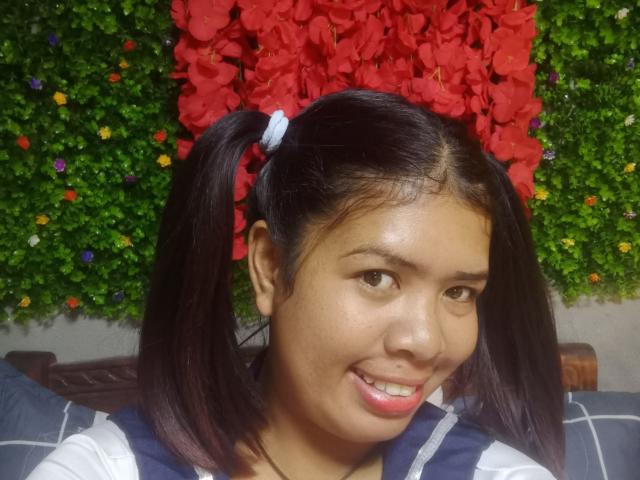 MariahSquirting69, una donna asiatica con guance paffute e trecce scure, sorride con le labbra rosse, indossa una camicia a righe e si trova di fronte a un muro fiorito verde e rosso..