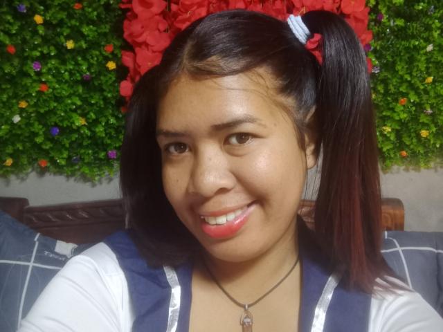 MariahSquirting69, Asian girl with black pigtails, wearing a red flower headband, smiling with pink lipstick, white and blue top, necklace with a heart charm, green leafy background.