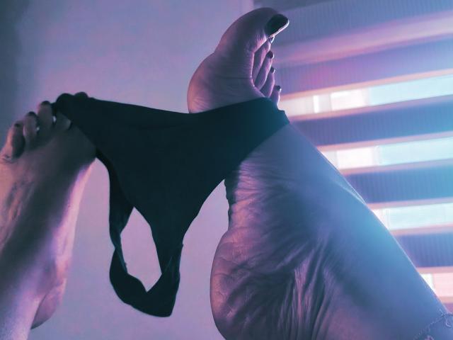 Il piede di Khharha è sollevato, e con le dita tiene delle mutandine nere, mostrando la pianta del piede e le unghie dipinte. Un'illuminazione soffusa in tonalità di viola e blu crea un'atmosfera sensuale e intima..
