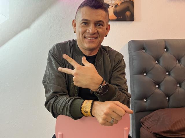 Cesirry, avec ses courts cheveux violets, portant une veste en cuir vert, une chemise noire et un bracelet doré, fait un signe de paix et pointe du doigt avec un sourire, assise devant un lit capitonné..