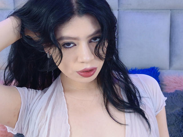 LindaMancini with black wavy hair, pale skin, and red lipstick, wearing a sheer white top, looking down with a slight pout, hand in hair, blue and pink fuzzy background.