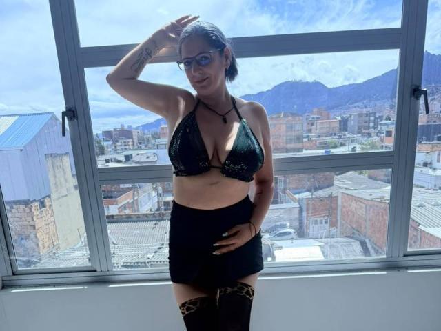MadameScarlett with glasses, black lace bra, big boobs, black mini-skirt, leopard thigh-highs, hand on hip, standing by window, mountains and city in background, sunny, tattoo on arm.