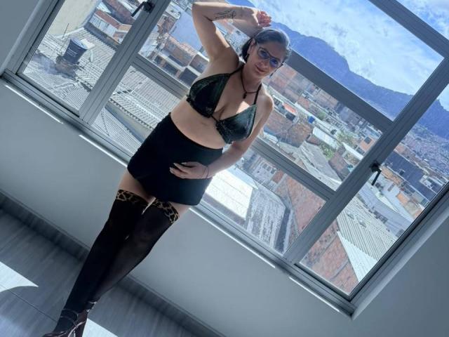 MadameScarlett stands in sunlight, wearing a black lace bralette, black skirt, and thigh-high stockings. Her tits are small, she has a tattoo on her left arm, and she's looking at the camera.