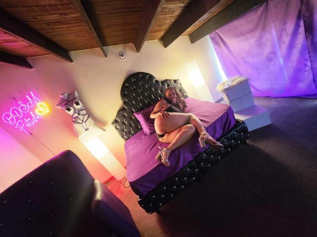 AnnyTanny, avec de grosses fesses et de petites seins, nue, sur un lit violet, les genoux repliés contre la poitrine, une tête de lit noire avec des touffes, un panneau lumineux au néon, un plafond en bois, un sol recouvert de moquette, des tiroirs blancs, des talons hauts..