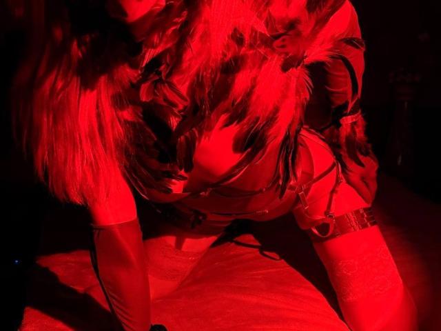 Fotografia con illuminazione rossa di MiraGi, con lunghi capelli ondulati, che indossa una giacca di pelle e un laccio, inginocchiata, che mostra il suo seno abbondante, con una visibile fascia sulla coscia e un guanto al braccio..
