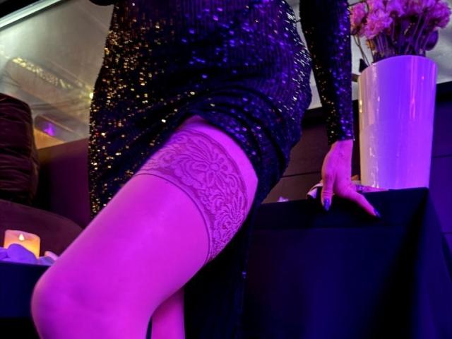 MiraGi, vestida con un elegante vestido negro, mostrando su trasero con medias altas de encaje, apoyada en una mesa con iluminación púrpura. Su mano descansa sobre la mesa..