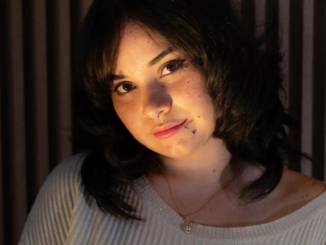AmaRavell, con i capelli neri ondulati, il viso cosparso di lentiggini e un sorriso delicato, indossa una maglietta bianca a coste e una collana dorata, illuminata delicatamente da un lato..