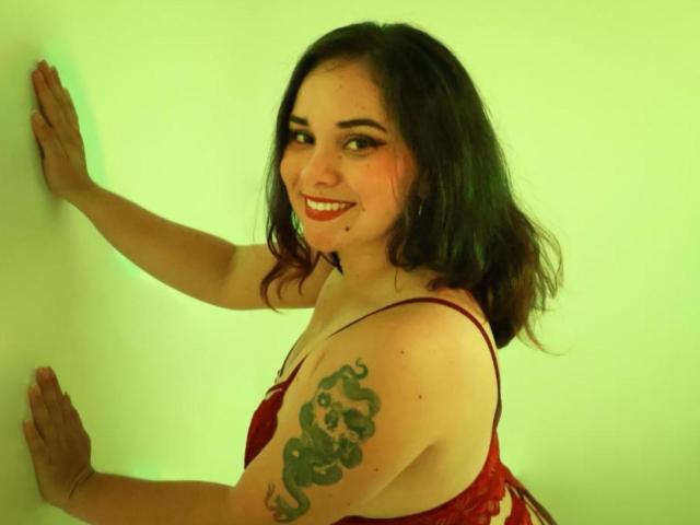 AmaRavell, avec ses cheveux noirs et ondulés, son rouge à lèvres rouge et son tatouage de serpent sur son bras droit, sourit tout en touchant un mur illuminé de vert avec les deux mains. Elle porte un soutien-gorge en dentelle rouge..