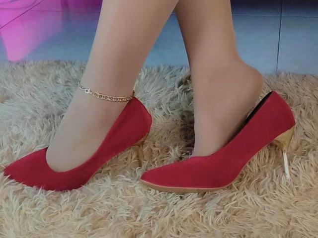 Vittoria, les jambes chaussées de chaussures à talons rouges aiguilles, avec une cheville ornée d'une chaîne dorée, se tient sur un tapis beige moelleux. Ses pieds sont pointus, mettant en valeur les talons hauts..