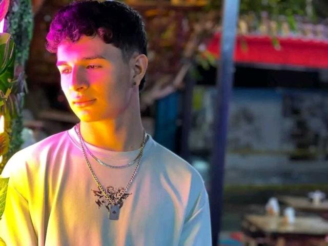 ValeriusAngel with short curly dark hair, wearing a white tee and two necklaces, standing in colorful, purple-lit light, looking to the side, with a subtle, calm expression.