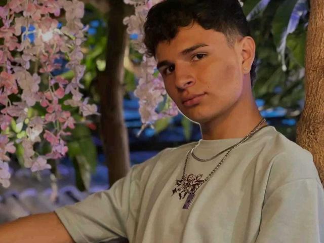 ValeriusAngel with curly brown hair, light skin, and a gold cross necklace, wearing a gray shirt, standing under pink flowers, looking at the camera with a slight smile.