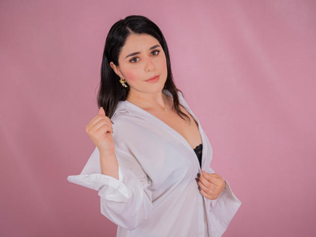VickyMegan with dark hair, pink cheeks, wearing a white shirt, black bra, gold earrings, and a necklace, slightly pulling her shirt, looking at the camera, smiling.