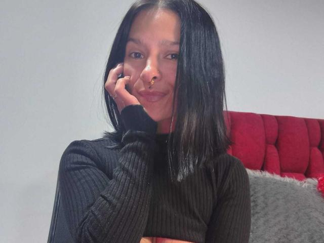 Amelie Blake, avec ses cheveux noirs lisses, son pull noir côtelé, son anneau de nez et sa main près de son visage, souriant légèrement. Un repose-tête rouge rembourré et un oreiller gris sont visibles en arrière-plan..