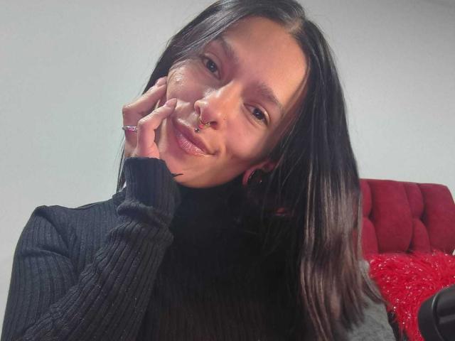 Amelie Blake mit schwarzen Haaren, Nasenring und Ohrringen, lächelnd, während sie ihre Wange auf ihre Hand legt, trägt einen schwarzen Rippstrick-Rollkragenpullover. Im Hintergrund ein roter Stuhl und ein Kissen..
