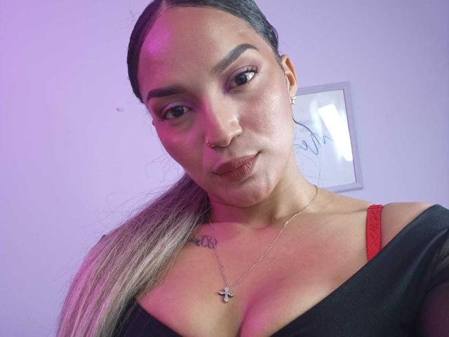 BrianaPratt, avec un anneau au nez, un haut noir laissant apparaître une bretelle de soutien-gorge rouge, de longs cheveux bruns, un tatouage discret sur l'épaule droite, portant un collier avec une croix, regardant la caméra avec un léger sourire..