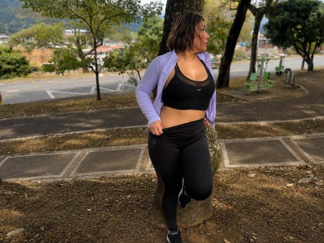 EmaFoxy, con cabello corto y oscuro, vestida con una camisa morada, un top deportivo negro y leggings negros, de pie, con una pierna levantada, mostrando sus grandes senos y sus curvas en un parque..