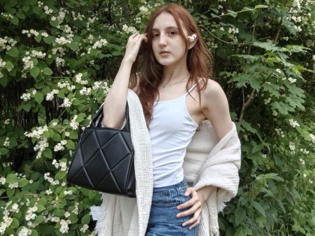 ReneUnderdown heeft rood haar, een wit tanktopje dat kleine borsten laat zien, blauwe jeans en een zwarte, gewatteerde tas. Ze draagt een witte cardigan en staat voor een groene achtergrond..