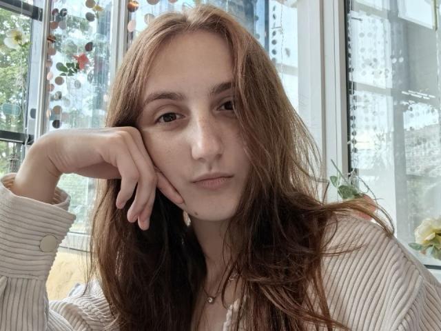 Rene Underdown tiene el pelo largo y castaño, pecas, y lleva una camisa beige texturizada. Tiene la cabeza apoyada en la mano, con una expresión seria y un ligero ceño fruncido, cerca de una ventana..