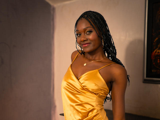 Andrea, peau foncée, longs cheveux tressés, robe en satin jaune, petit collier en or, souriant à la caméra, montrant ses seins, debout dans une pièce éclairée doucement, avec un tableau sur la droite..